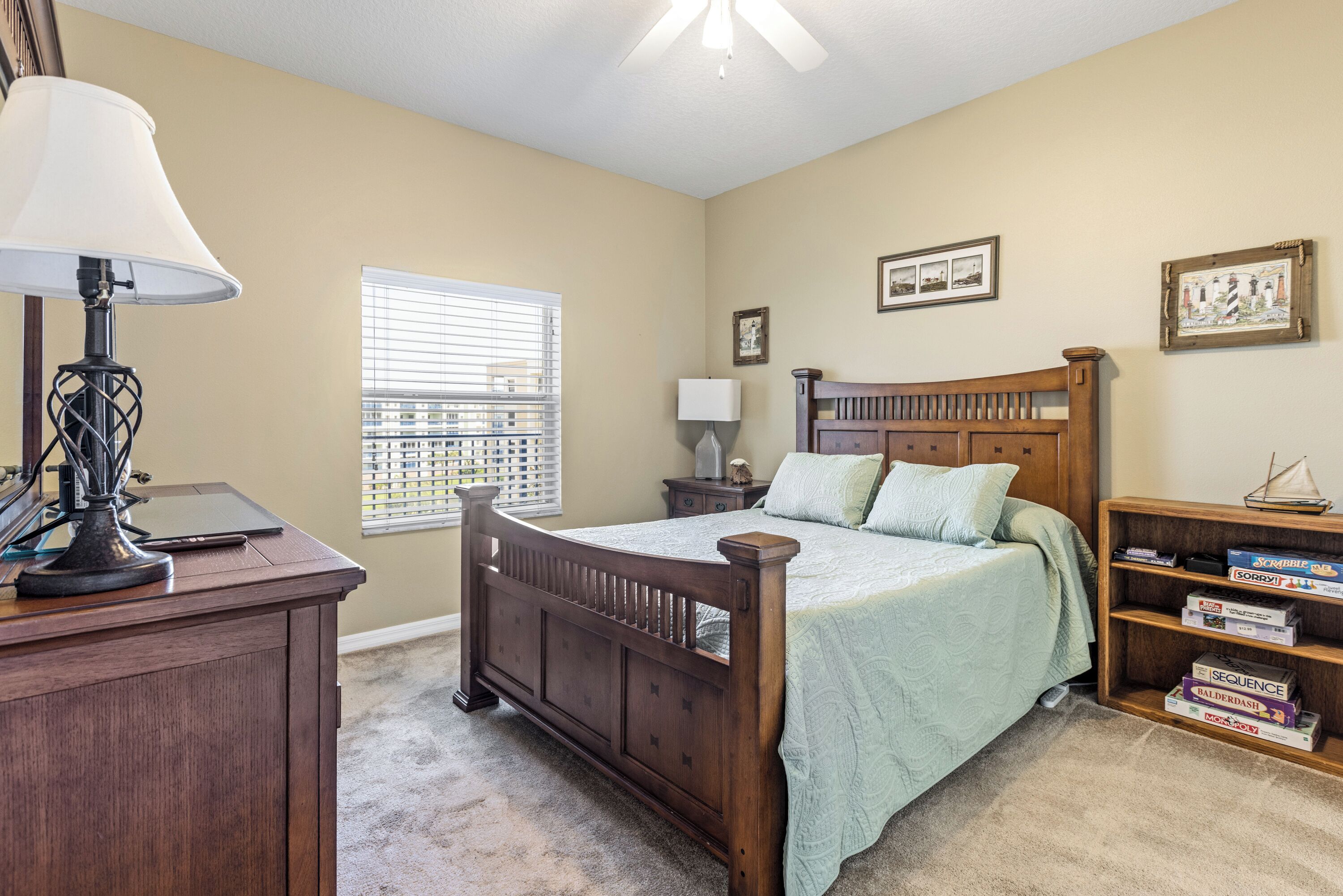 A bedroom with a wooden bed, a desk with a lamp, a window with blinds, and a bookshelf with board games.