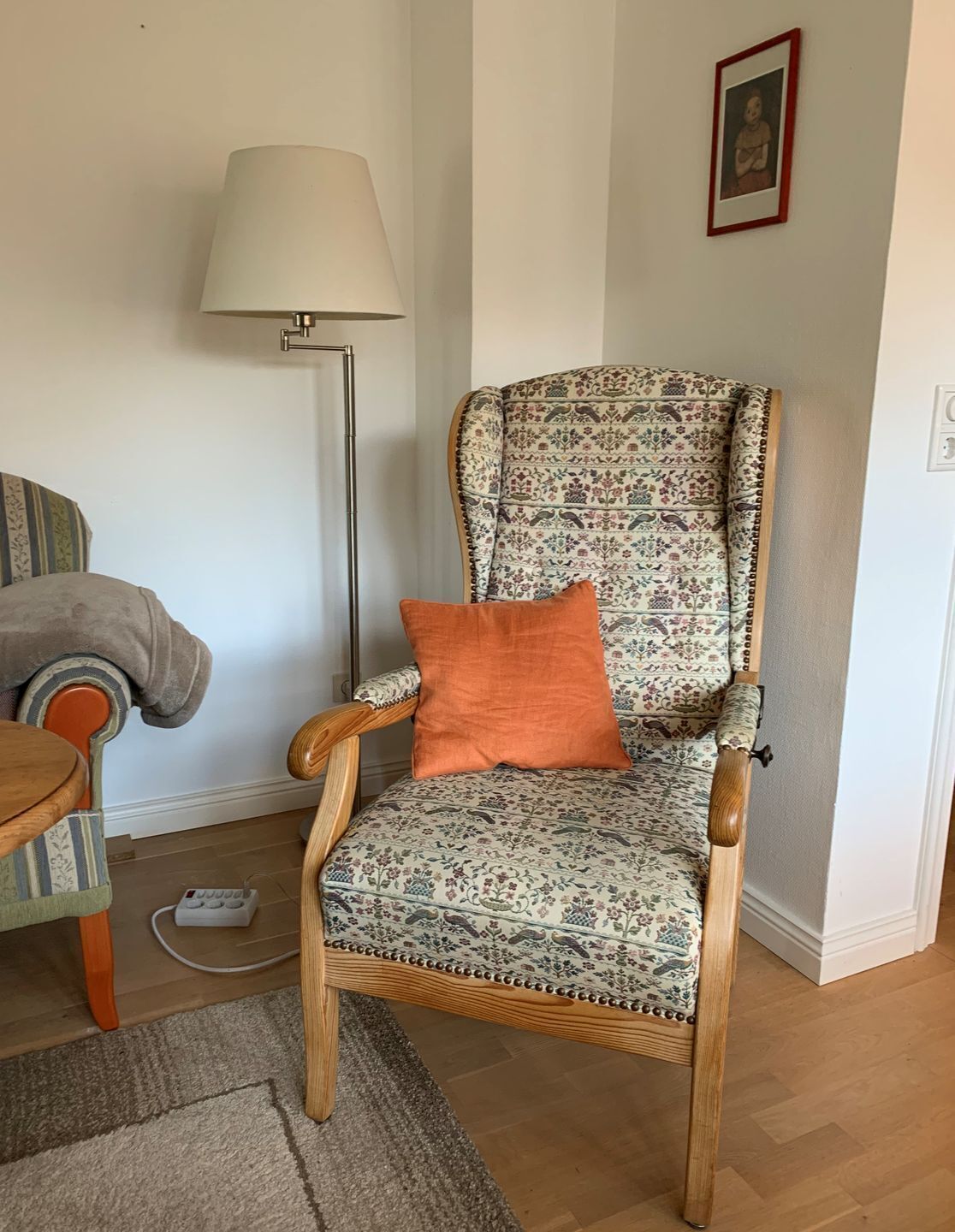 Ein Holzschaukelstuhl mit floralem Stoffbezug und einem orangefarbenen Kissen, platziert in einem Raum mit Stehlampe und einem gerahmten Bild an der Wand.