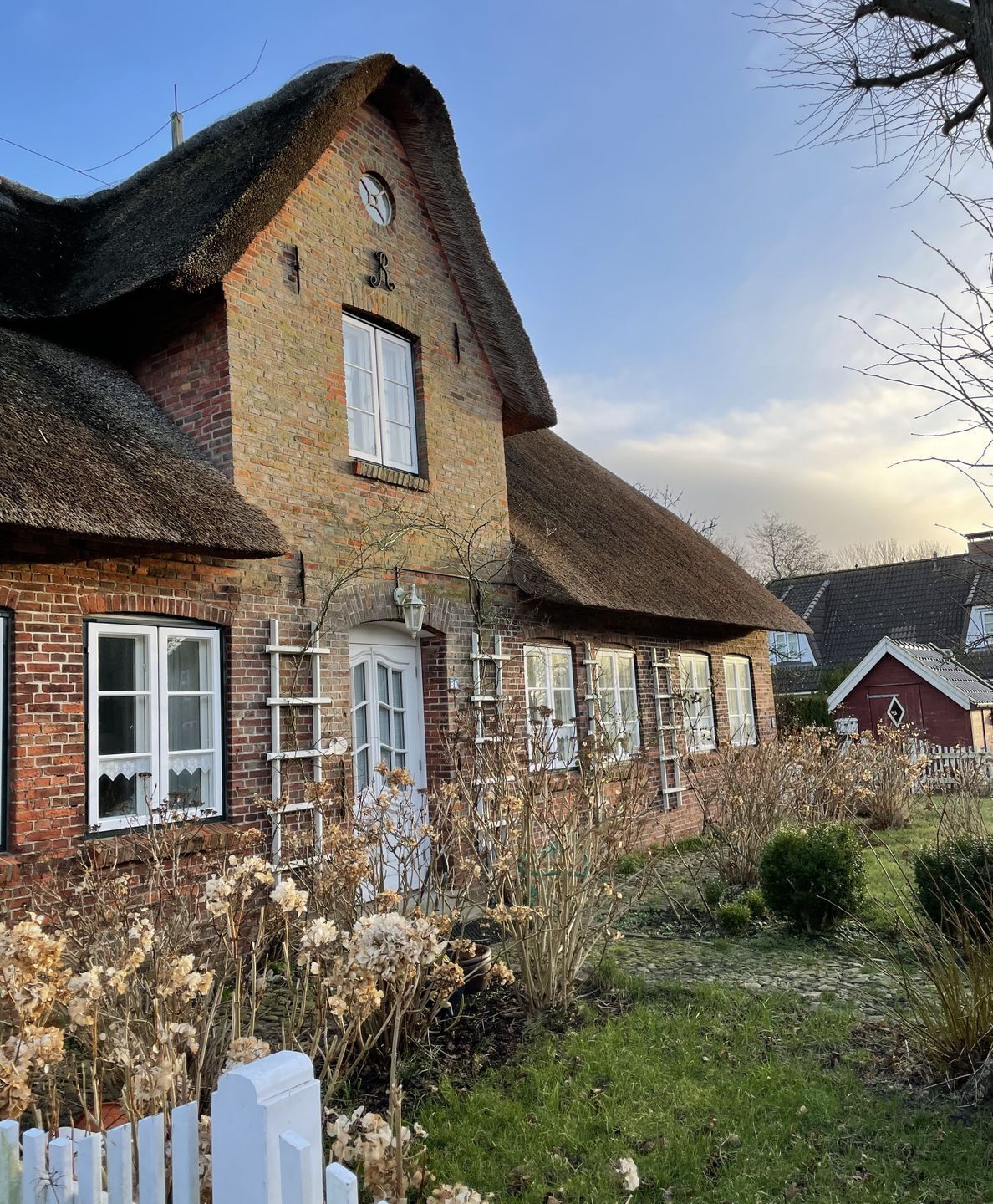 A traditional brick house with a thatched roof, a white picket fence, and a garden with dried plants.