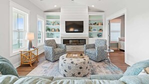 A living room with a fireplace, two patterned armchairs, a round ottoman, a television, and built-in shelving. - Carlton by Brunswick Vacation Rentals: Private Pool, Elevator, and Beach Access (Supply)