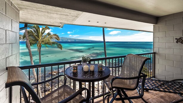 Un balcón con mesa y sillas, con vista a una playa con palmeras y agua azul cristalina.