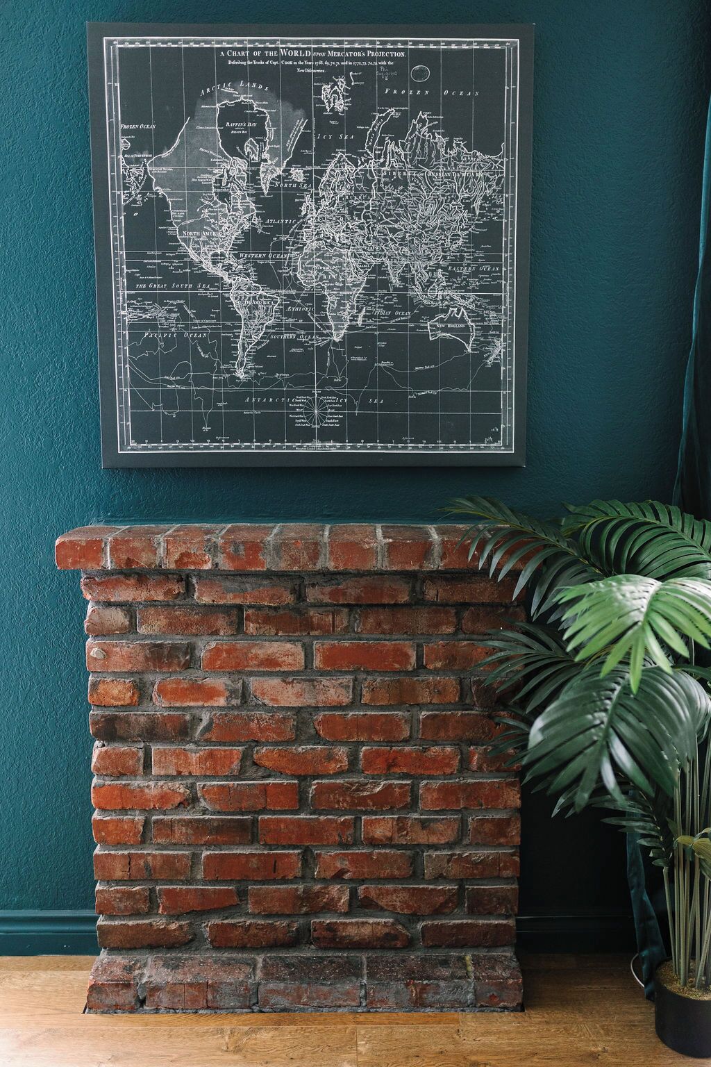 Un mapa mundial enmarcado sobre una pared verde azulada, encima de una chimenea de ladrillo.