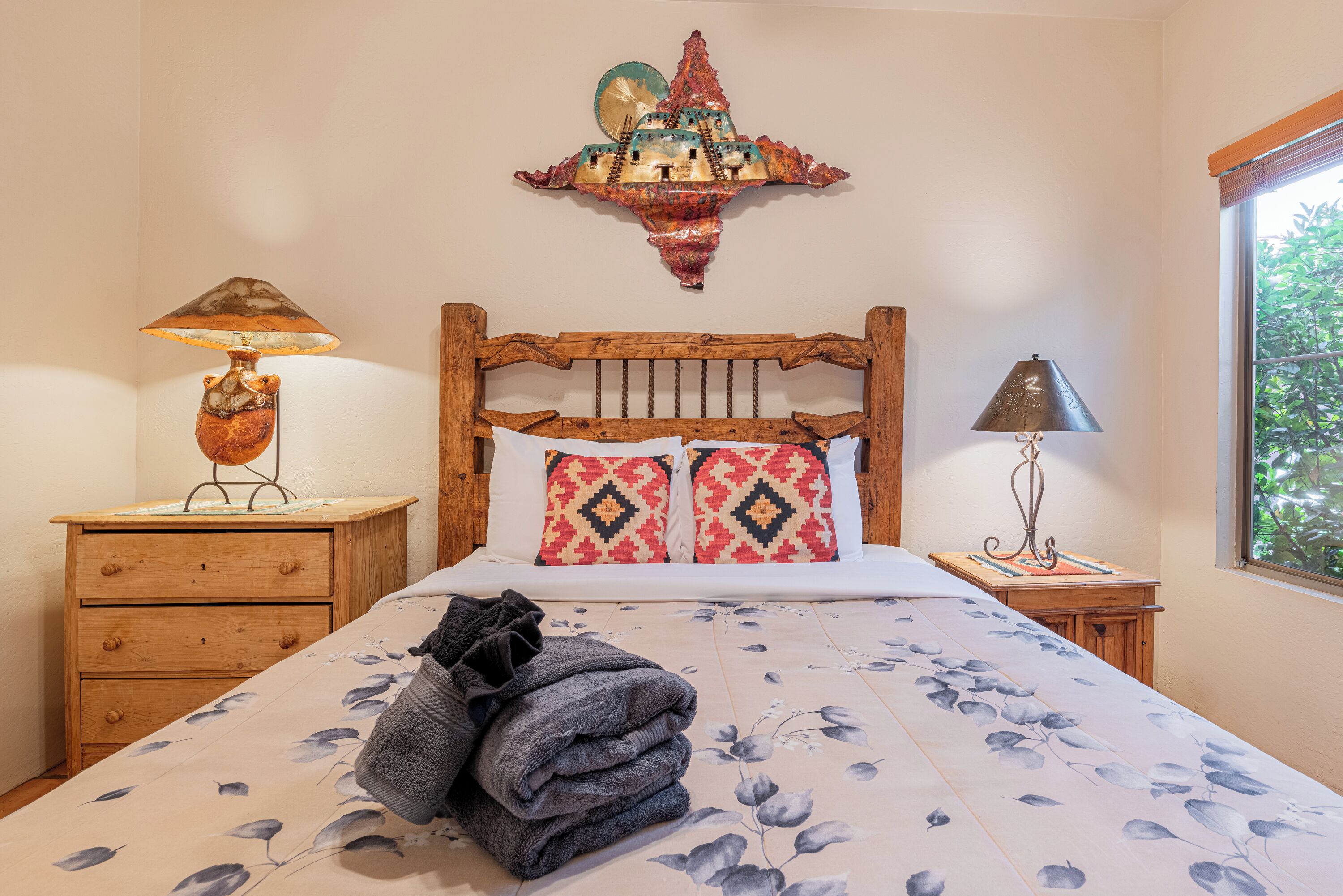 A bedroom with a wooden headboard, a bed with floral bedding, two patterned pillows, a nightstand with a lamp, and a decorative wall piece.