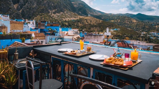 A rooftop terrace with a table set for breakfast, overlooking a mountainous landscape. - Riad Azurite (Chefchaouen)