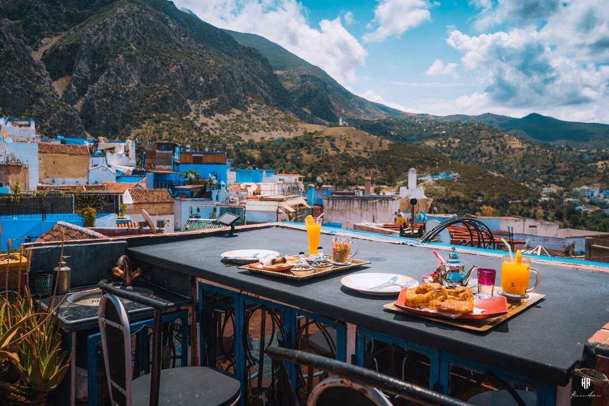 A rooftop terrace with a table set for breakfast, overlooking a mountainous landscape.