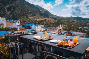 A rooftop terrace with a table set for breakfast, overlooking a mountainous landscape. - Riad Azurite (Chefchaouen)