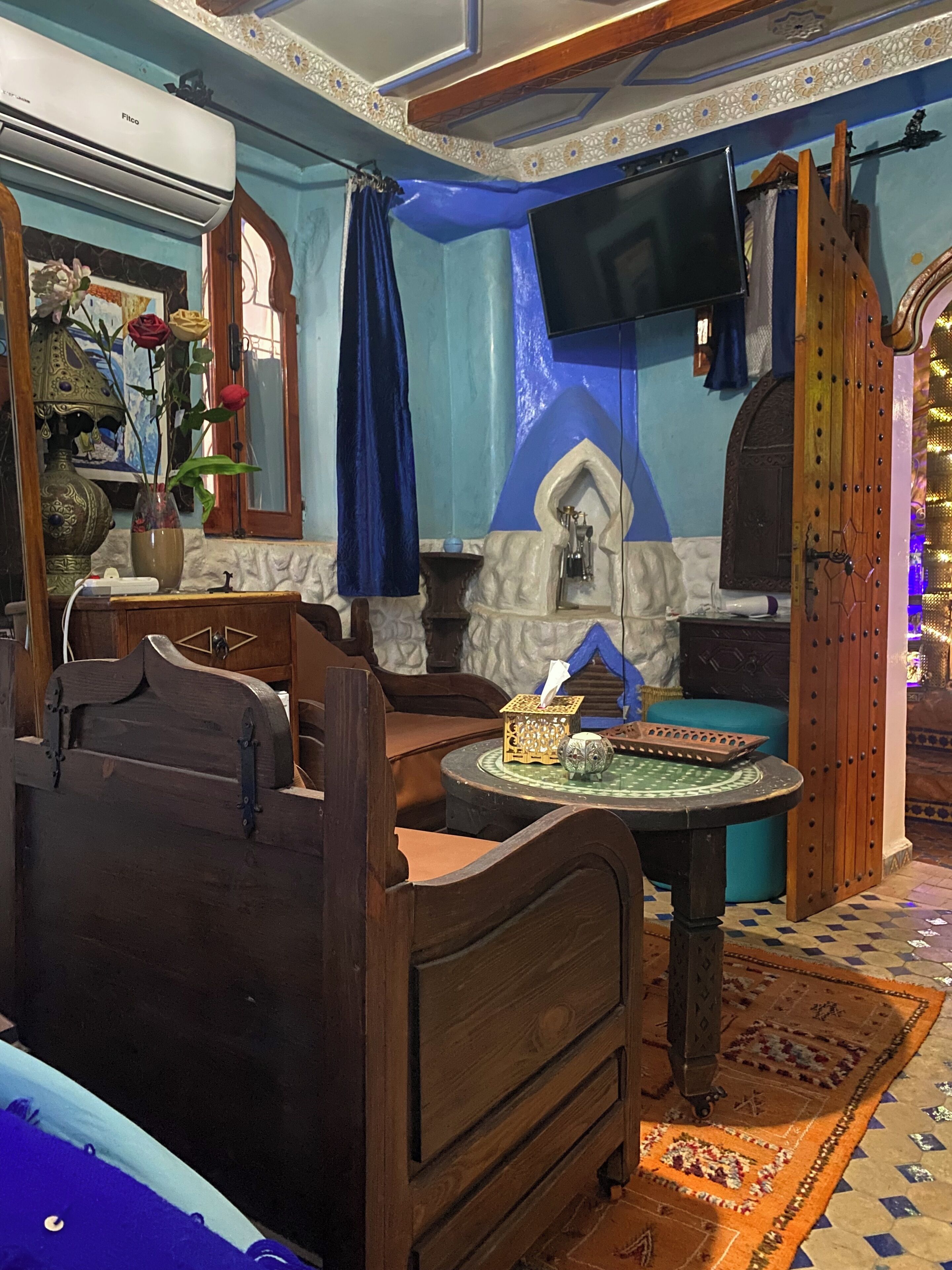 A traditional room with a bed, a wooden chest, a small table with a decorative item, a wall-mounted TV, and a blue curtain.