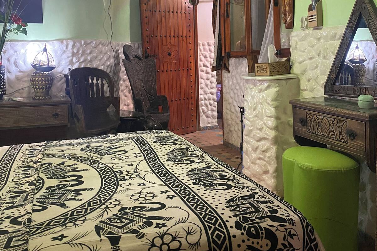 Quarto com cama, guarda-roupa de madeira, um banquinho verde, televisão fixada na parede e um espelho decorativo.