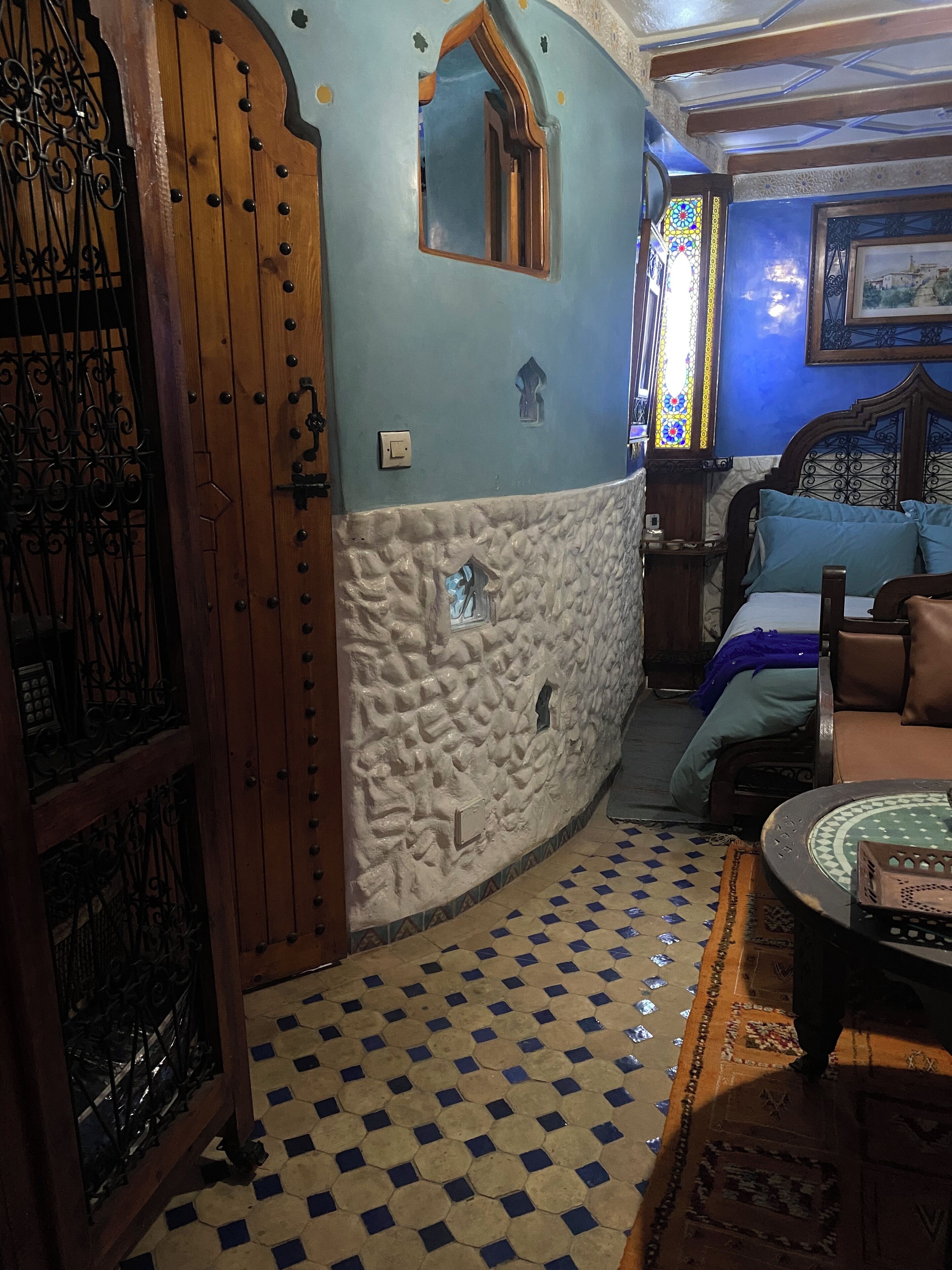 A room with a stone wall, a wooden door with metalwork, a bed with blue bedding, and a patterned floor.