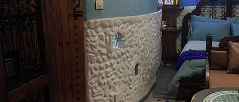 A room with a stone wall, a wooden door with metalwork, a bed with blue bedding, and a patterned floor.