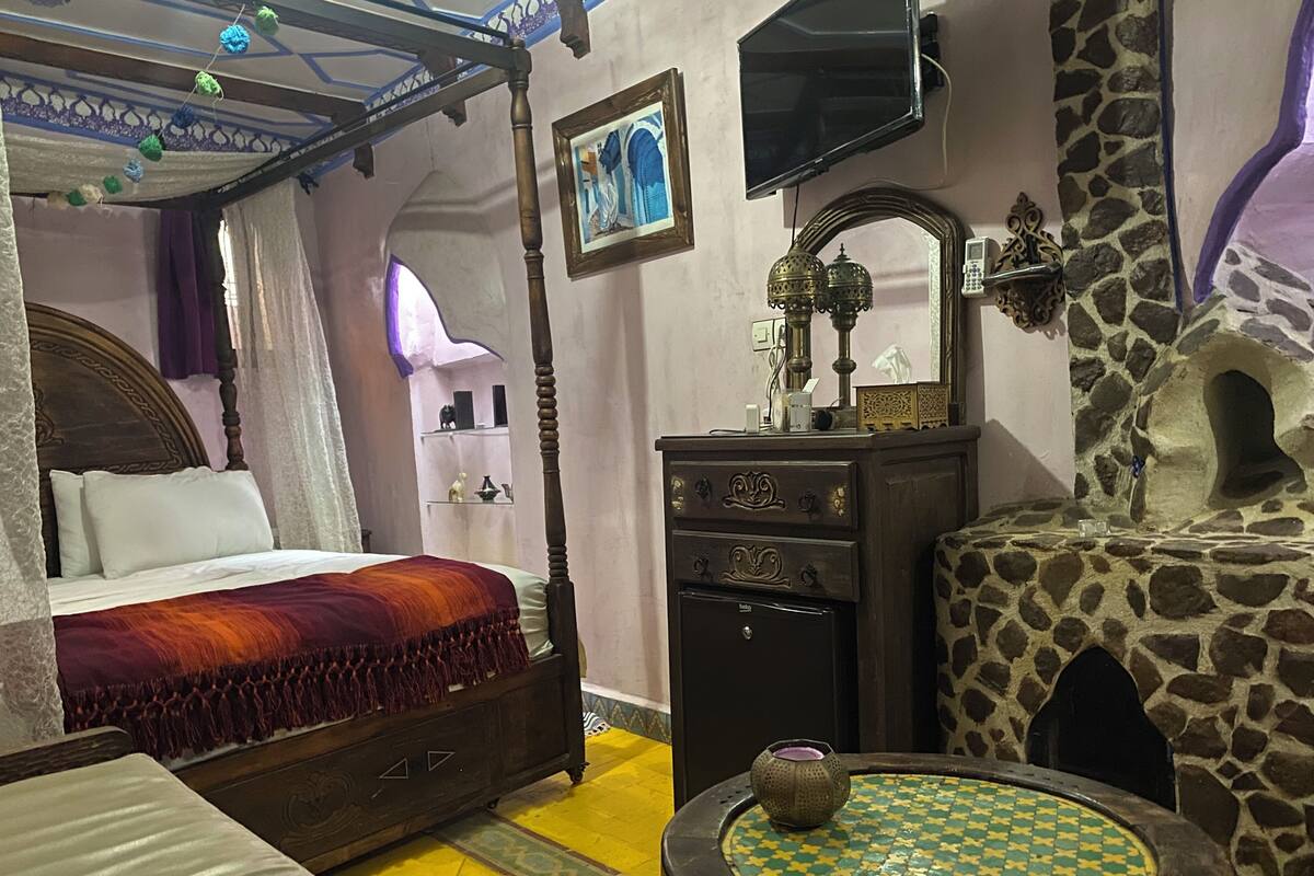 Quarto tradicional com uma cama de dossel, um painel de parede de pedra, uma pequena mesa redonda e uma televisão fixada na parede.