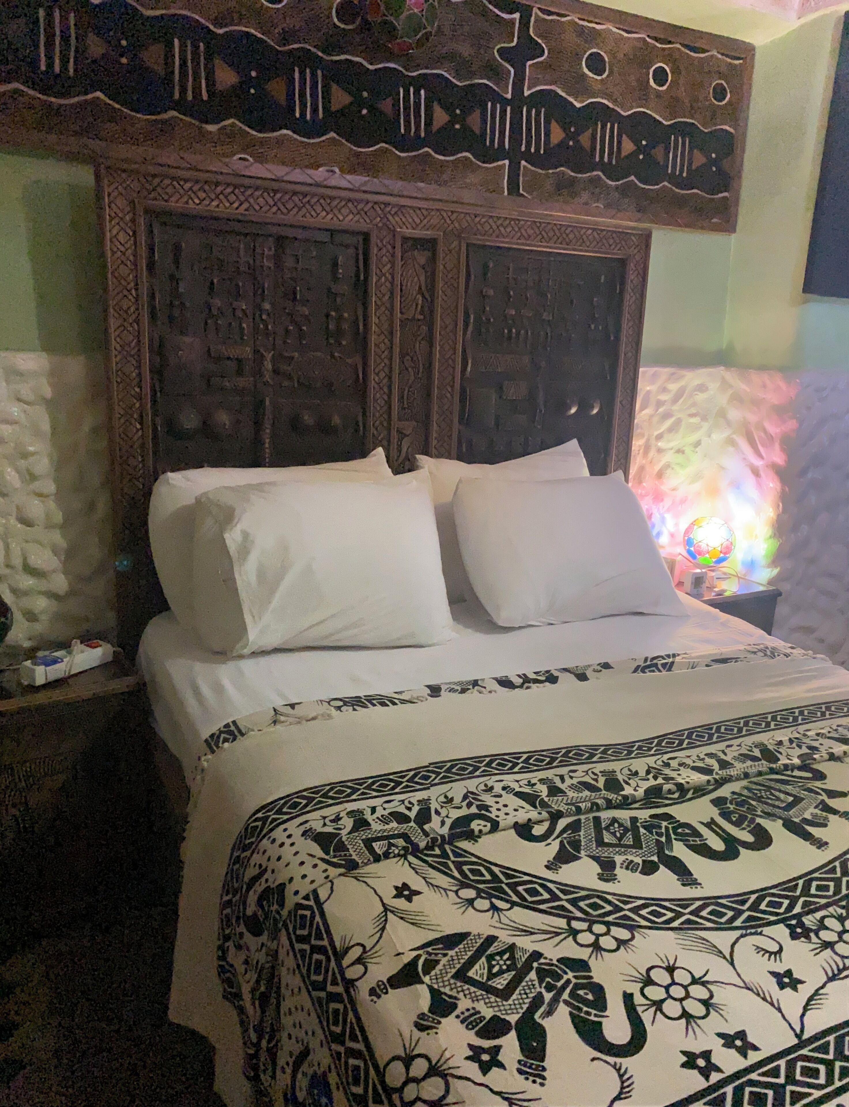 Ein Bett mit weißen Laken und einer gemusterten Tagesdecke, ein Holz-Kopfstück mit aufwendigen Schnitzereien und eine Wand mit einer Zierleiste.