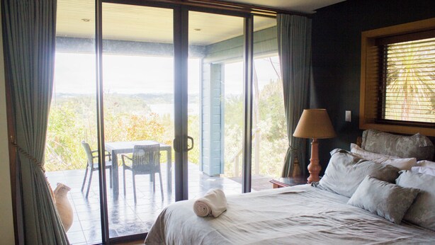 Une chambre avec un lit, une table de chevet, une lampe, une fenêtre avec des stores et une vue sur un patio équipé d’une table et de chaises.