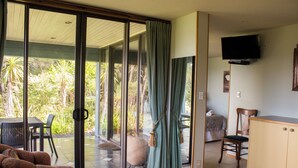 Une pièce dotée d’une porte-fenêtre coulissante donnant sur une terrasse avec vue sur la végétation tropicale. On y trouve un canapé, un fauteuil, un meuble et un téléviseur fixé au mur.