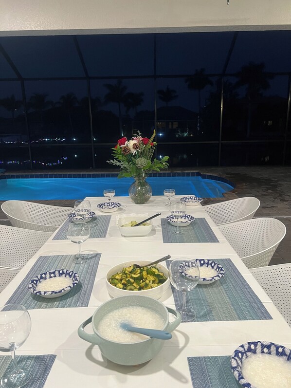 An outdoor dining area with a pool view, white wicker chairs, and a table set with plates, bowls, and a vase of flowers.