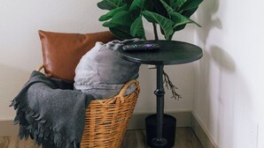 Een hoek van een kamer met een rieten mand, een bruin kussen, een grijze deken en een kunstplant in een groene pot op een zwarte tafel.