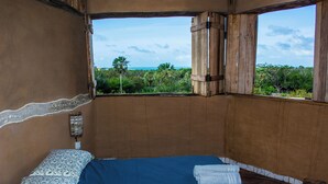 Une chambre avec un lit, une fenêtre donnant sur la plage et les palmiers, et une porte en bois.