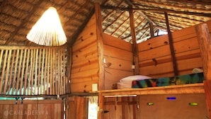 A wooden cabin interior with a thatched roof, a bed with a colorful blanket, and a hanging light fixture. - Raízes Eco Hostel (Cajueiro da Praia)