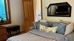 A bedroom with a bed, a wooden dresser, a desk, a television mounted on the wall, and a window with blinds.