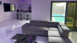 A modern living room with a grey sectional sofa, a glass coffee table, and a kitchen area in the background. - Tino Luxury House & Residence (Mbour)