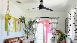 Ein Wohnzimmer mit braunem Ledersofa, einem Couchtisch, einem Fernseher und einem Deckenventilator. Ein großes Fenster mit Vorhängen, eine Pflanze und eine Tür nach draußen sind ebenfalls vorhanden.