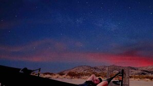 Eine Person liegt nachts in einer Hängematte an einem sandigen Strand, über ihr leuchtet ein Sternenhimmel.