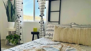 Ein Schlafzimmer mit einem Bett, Kissen, einer Decke, einer Zimmerpflanze und einem kleinen Tisch.