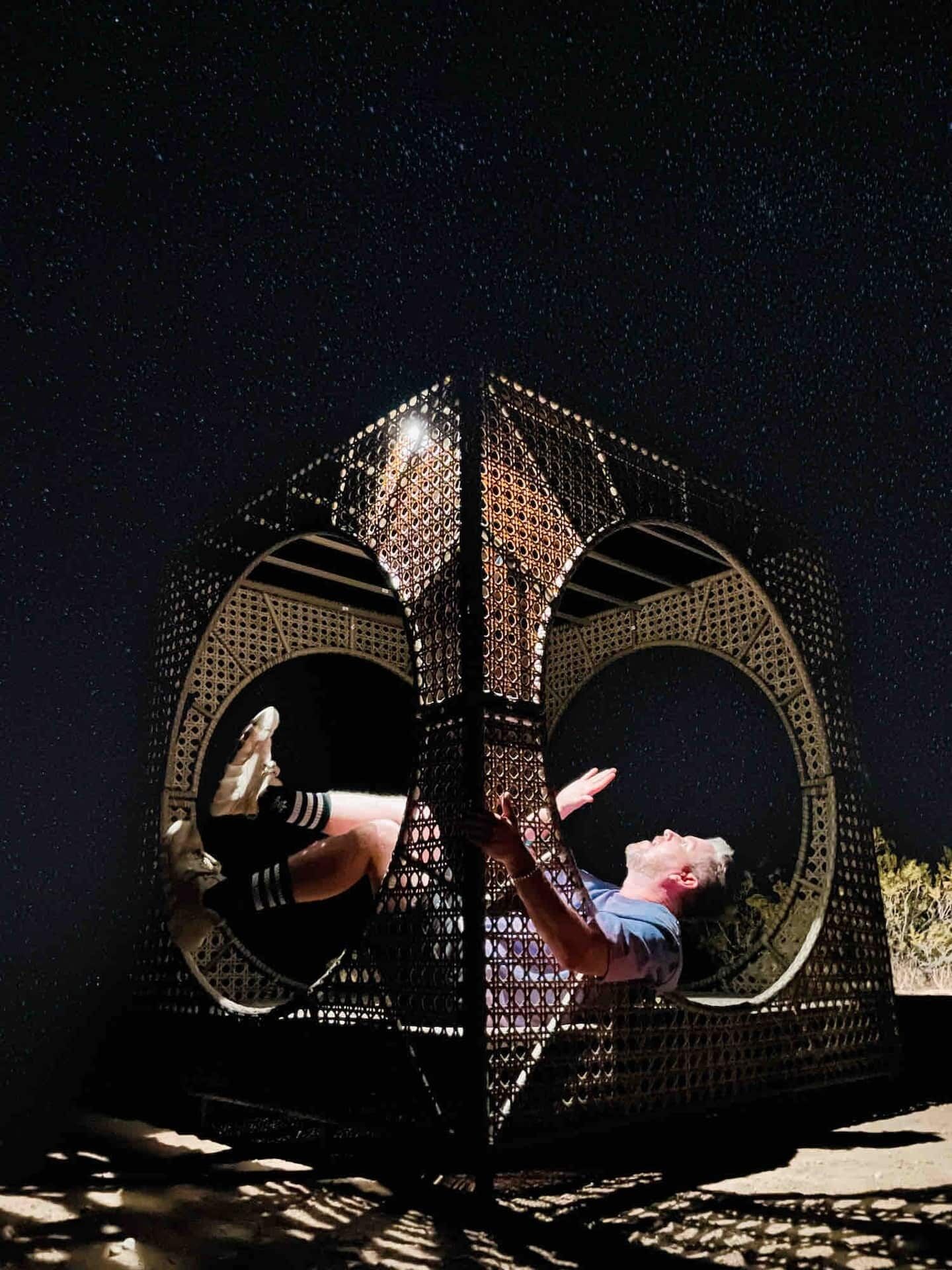 A person lying inside a large, circular, metallic structure with a starry night sky in the background.