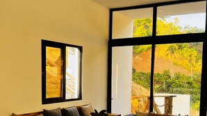 A modern dining area with a wooden table set for four, a sofa, and a large window offering a view of greenery. -  10 min from the beach!Romantic Spanish apto w/Infinity Pool. (Las Terrenas)