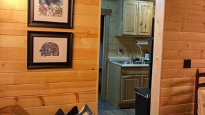 A cozy wooden cabin interior with a living area featuring a couch and pillows, framed artwork on the wall, and a view into the kitchen area with wooden cabinets and a sink. - Cozy pet friendly cabin with stand alone sauna near Willow and Talkeetna Alaska (Willow)