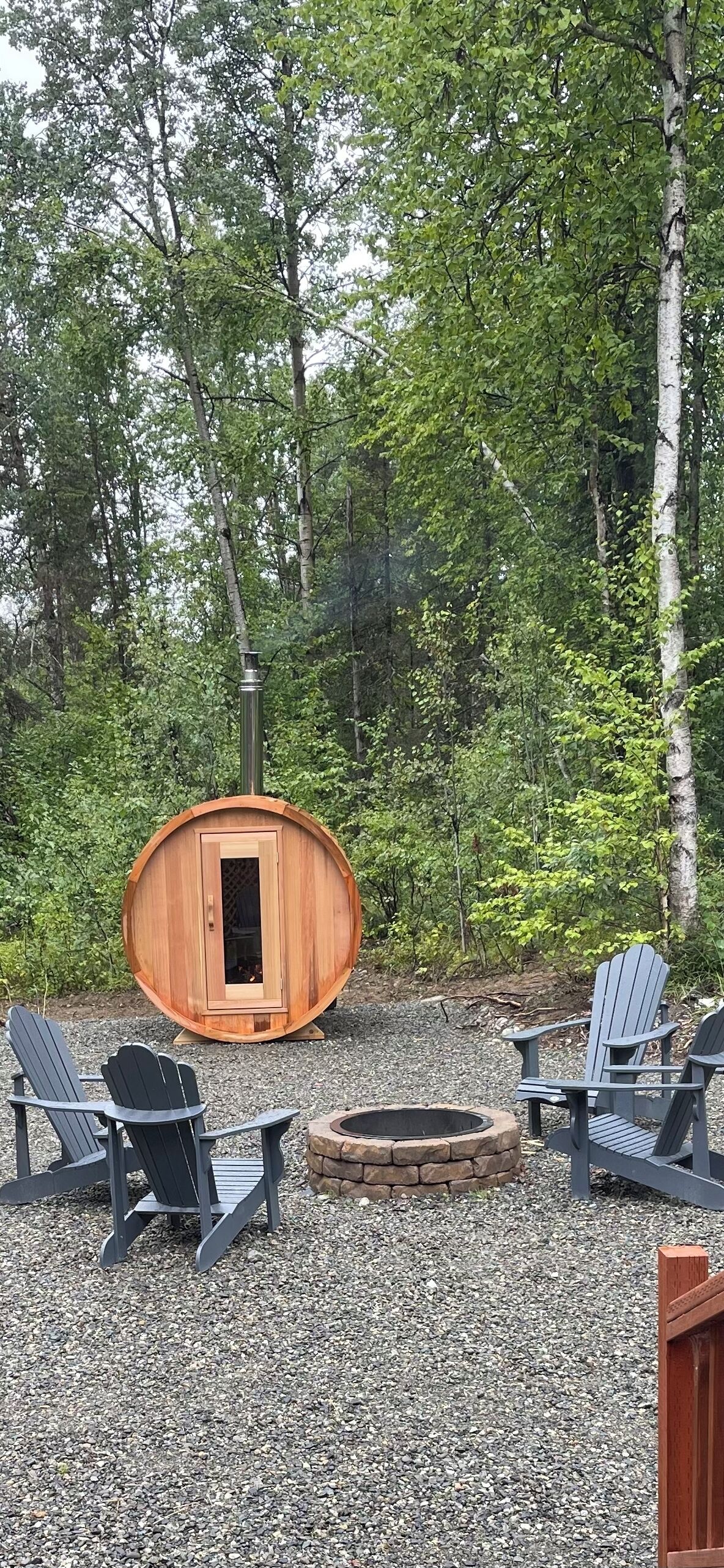 Eine Holzsauna im Wald mit zwei Adirondack-Stühlen und einer Feuerstelle.