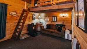 A cozy cabin interior with a loft bedroom, wooden walls, a fireplace, and a staircase. - Cozy pet friendly cabin with stand alone sauna near Willow and Talkeetna Alaska (Willow)