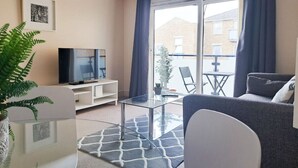Un salon moderne avec un téléviseur à écran plat, une table basse en verre, un canapé gris et une grande fenêtre avec des rideaux.