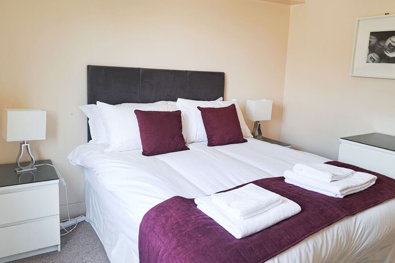 A neatly made bed with white linens and maroon pillows, a purple throw blanket, and two bedside lamps on white nightstands.