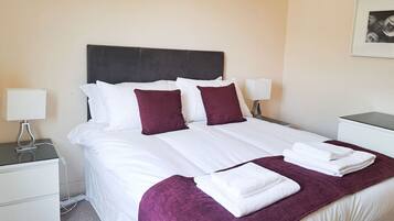 A neatly made bed with white linens and maroon pillows, a purple throw blanket, and two bedside lamps on white nightstands.