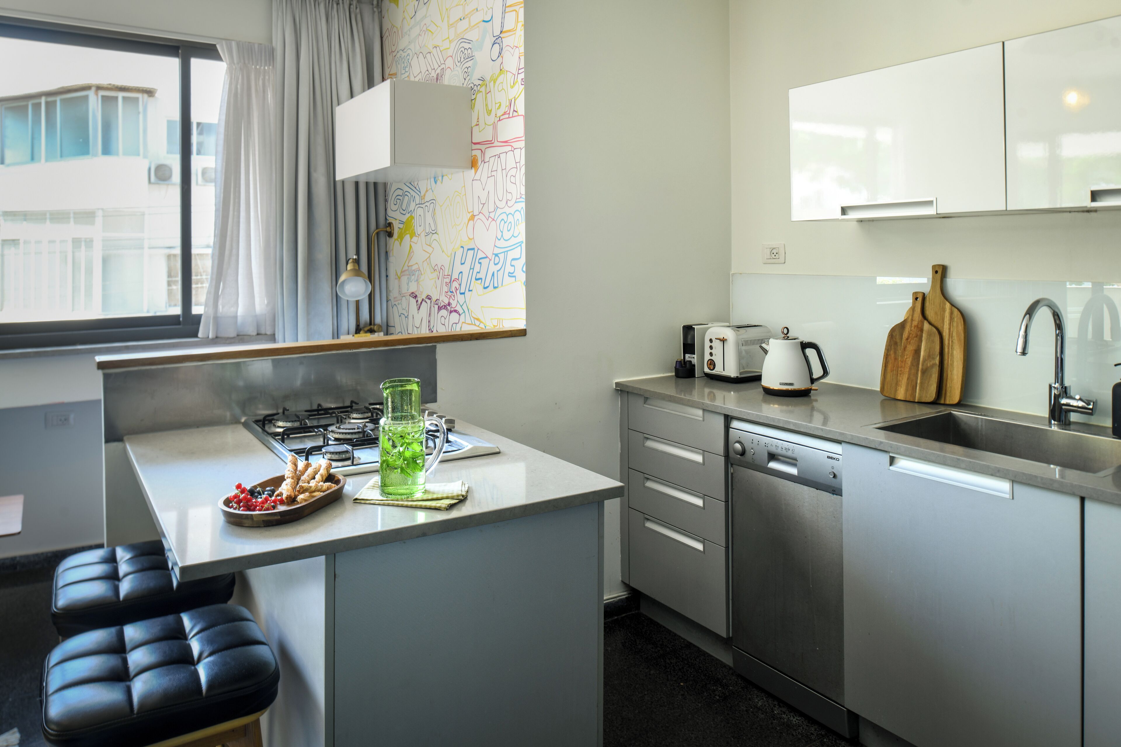 A modern kitchen with a gas stove, a dishwasher, and a sink. There is a window with curtains, a map on the wall, and a small seating area with a bench.