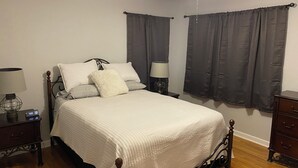 Ein ordentlich bezogenes Bett mit weißer Decke und Kissen, ein Nachttisch mit Lampe sowie ein Beistelltischchen mit einer Uhr.