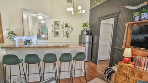 A modern kitchen with a bar area, green bar stools, a wooden cabinet with books, and a television on a wooden stand. - 2 Bdrm Sits on Riverwalk (San Antonio)