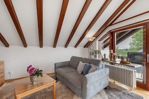Ein Wohnzimmer mit grauem Sofa, Holzbalken, Heizkörper und einem Fenster mit Ausblick.