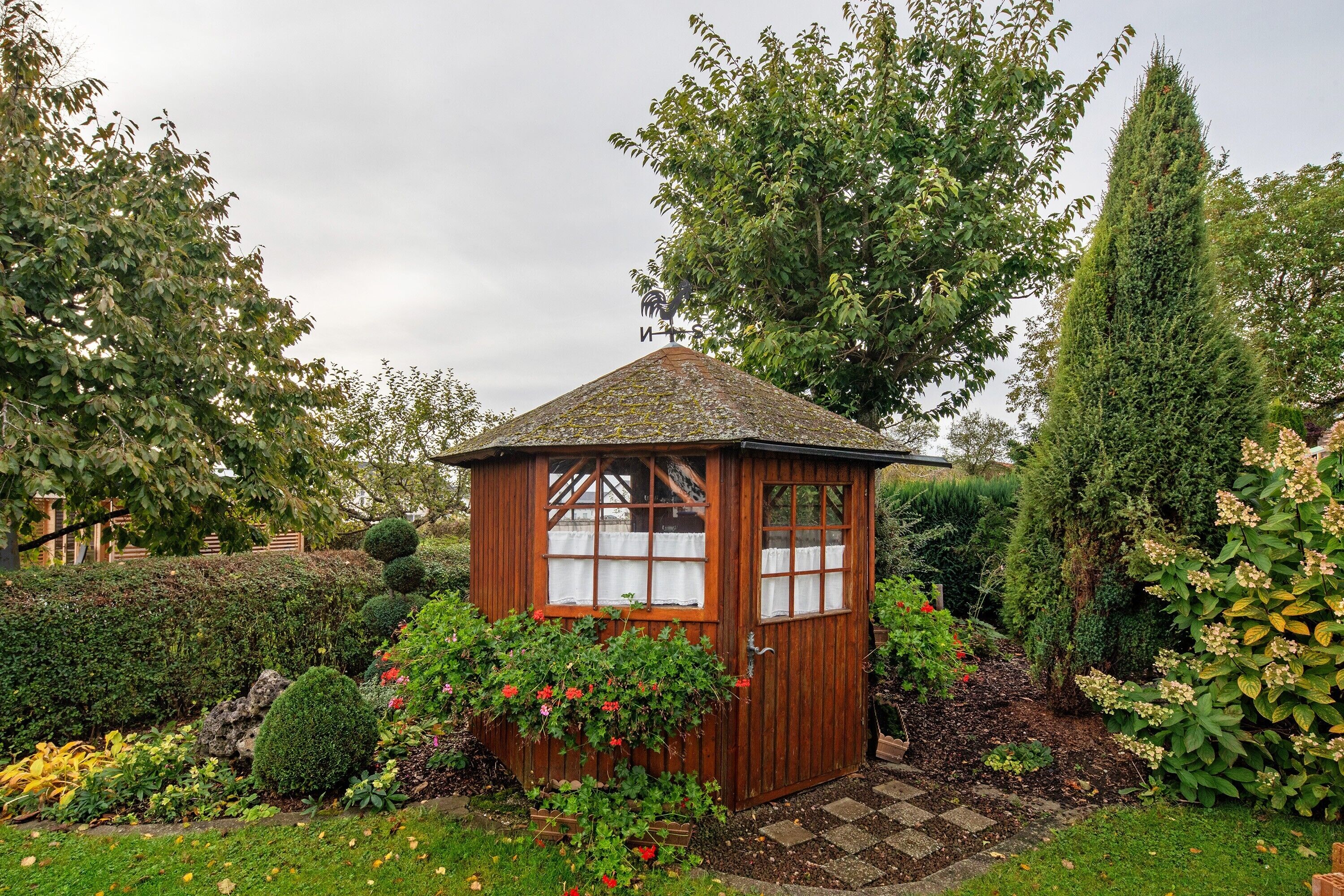 Un abri de jardin en bois, doté d’un toit en pente, entouré d’une végétation luxuriante et de plantes en fleurs.