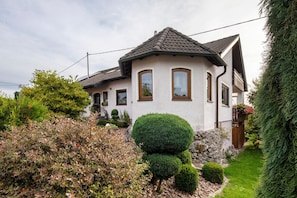Ein zweistöckiges Haus mit Natursteinfundament, einem Ziegeldach und einem gepflegten Garten mit verschiedenen Sträuchern und einer ordentlich geschnittenen Hecke.