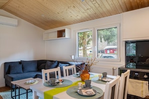 Uma sala de estar moderna com teto de madeira, um sofá, uma mesa de jantar com jogos americanos, um vaso com galhos e vista para o exterior através de grandes janelas.