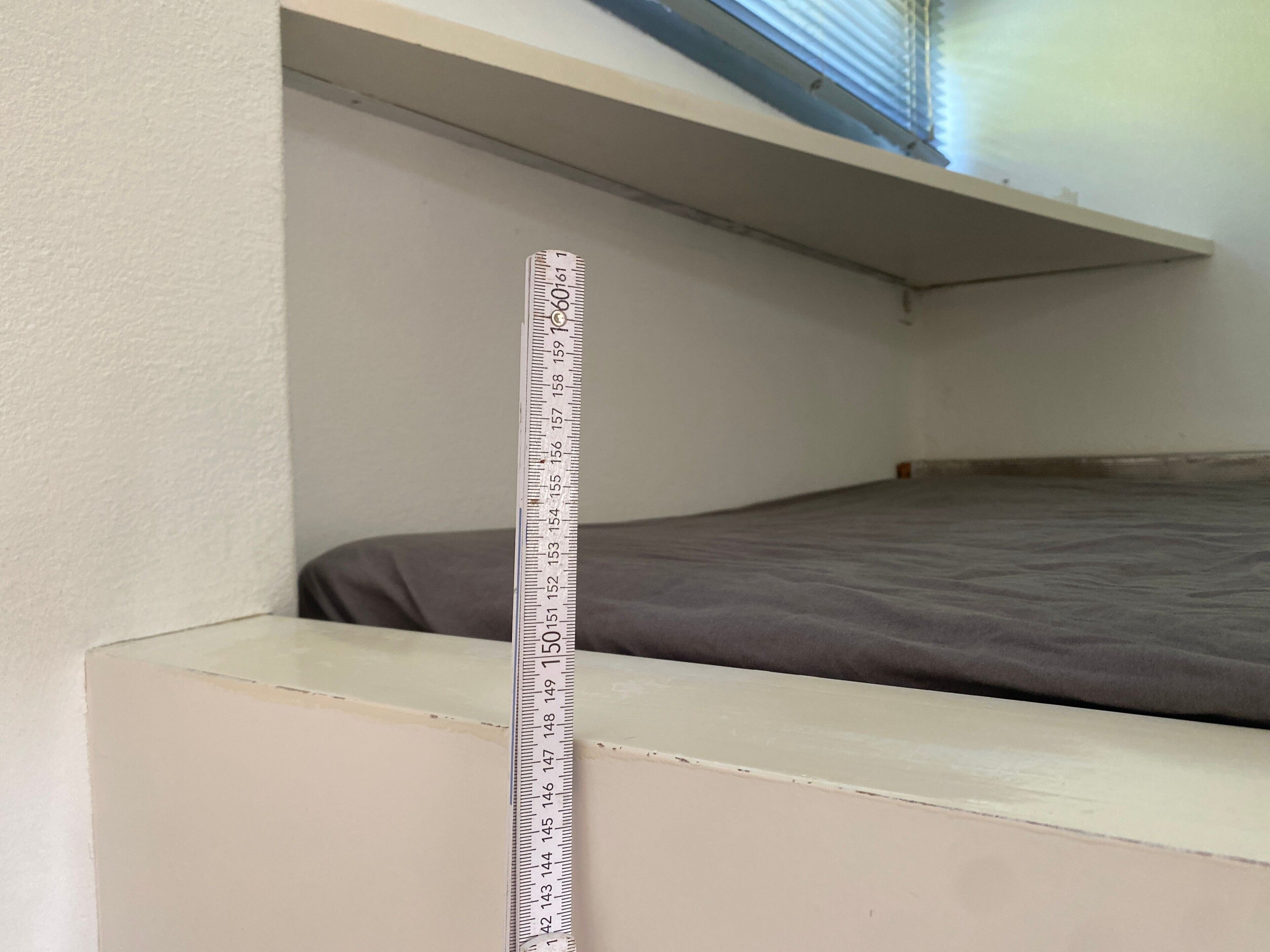 Ein Maßband an einer Wand, ein Bett und eine Decke mit einem Oberlicht.