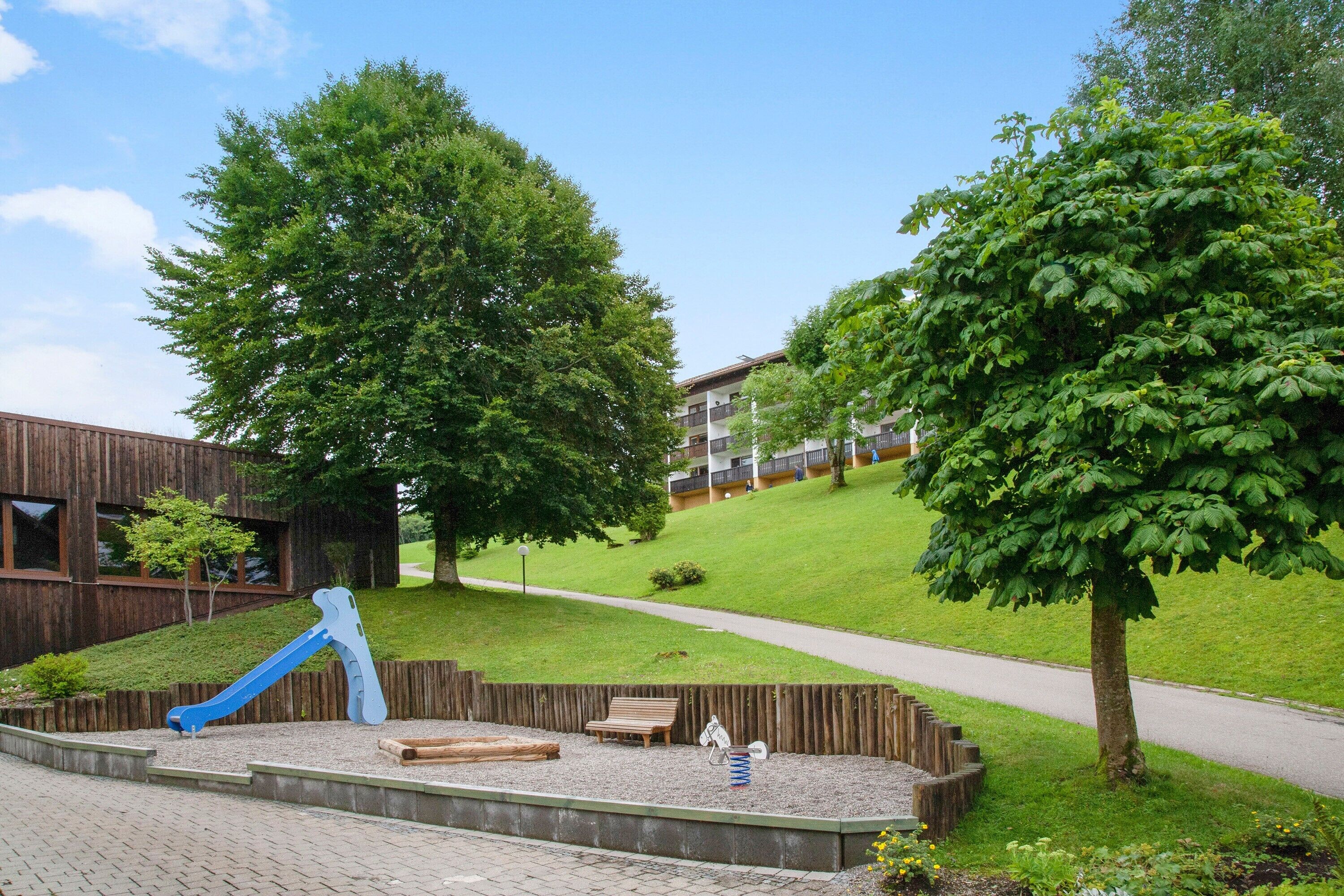 A playground with a blue slide and swings, surrounded by trees and a grassy hill.