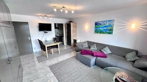 A modern living room with a grey sofa, a glass coffee table, a dining area with a table and chairs, a flat-screen TV mounted on the wall, and a painting of a landscape.