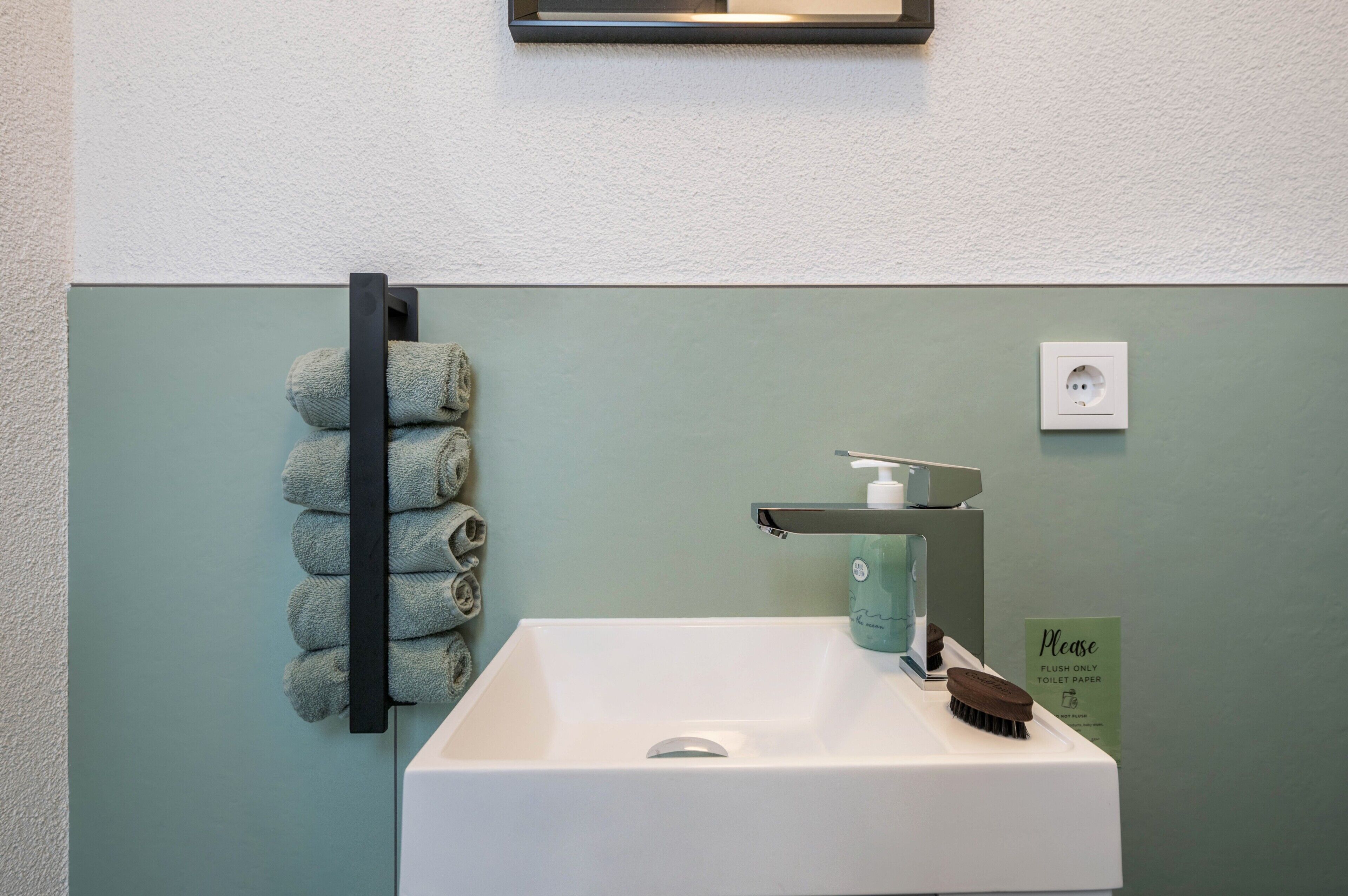 A bathroom with a white sink, a towel rack with rolled towels, a soap dispenser, and a flush-only toilet paper sign.
