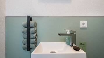 A bathroom with a white sink, a towel rack with rolled towels, a soap dispenser, and a flush-only toilet paper sign.