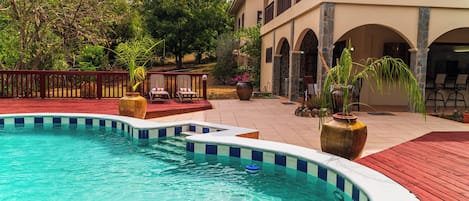 A swimming pool with a red deck, surrounded by potted plants and lounge chairs.