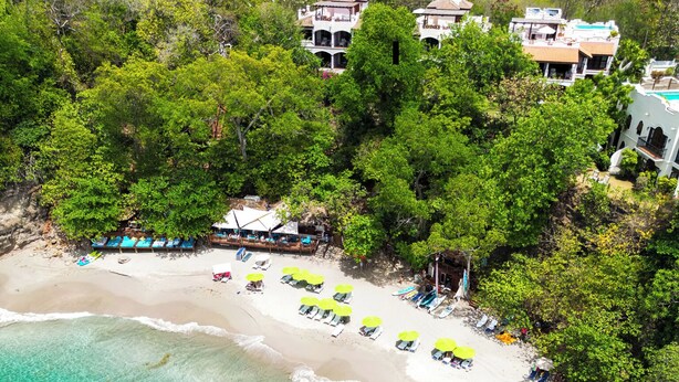 Une plage aux eaux turquoise et limpides, une rangée de transats et un bâtiment doté d’un balcon donnant sur la plage.
