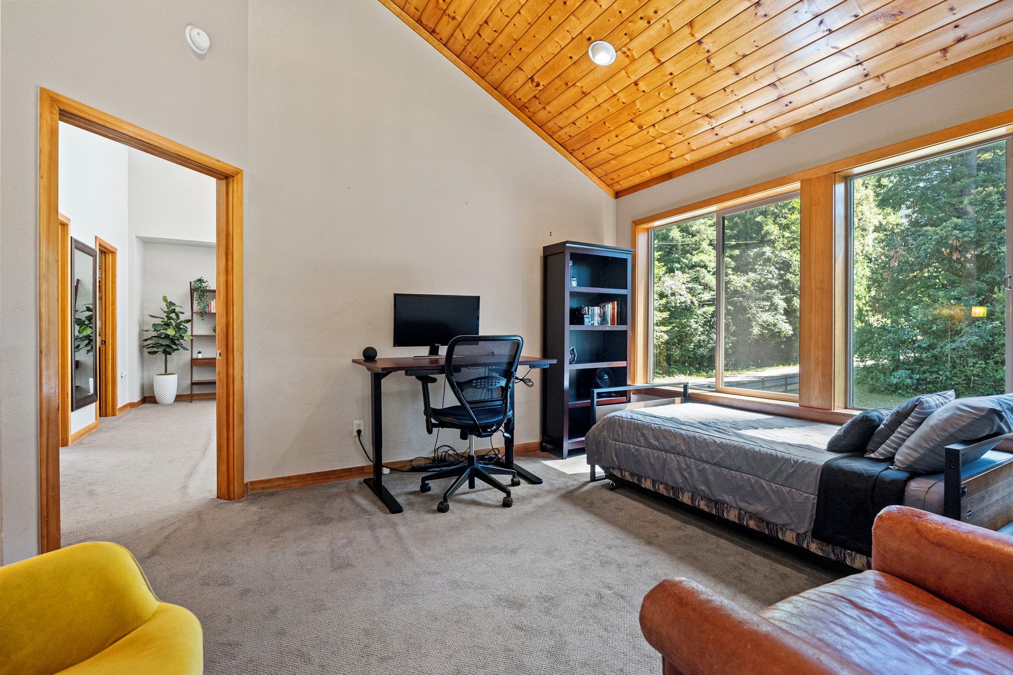 Un dormitorio con cama, escritorio con computadora, estantería y ventanas amplias.